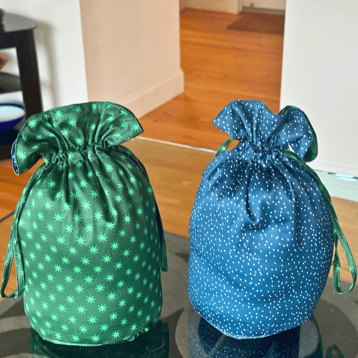 Two fabric drawstring reusable fabric gift bag pouches on a glass table in an indoor room. The left pouch is green with a light green star pattern, and the right pouch is blue with white polka dots.